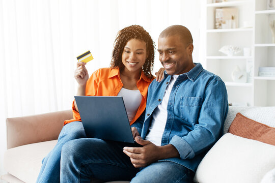 Online Shopping. Happy Black Couple With Laptop And Credit Card Relaxing On Couch At Home, Cheerful African American Spouses Making Easy Payments, Enjoying Internet Purchases, Copy Space