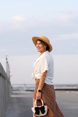 Smiling woman wearing straw hat walks outdoors at sunset.