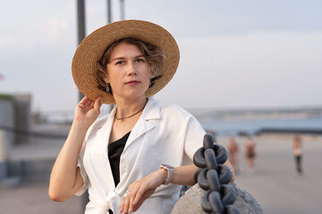 Elegant woman thoughtfully adjusts straw hat at seaside promenade.