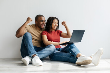 Joyful african american couple sitting on floor with laptop and celebrating success, happy black spouses raising arms in victory, cheering good news, posing over white wall background, free space