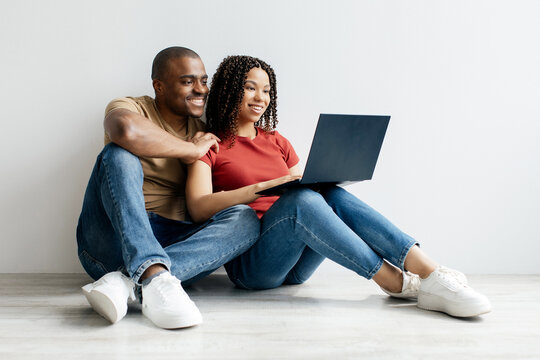 Young Black couple sitting on floor at home, smiling while using laptop together. African American spouses browsing interne, experiencing casual scene of bonding, relaxation, and shared moments - Powered by Adobe