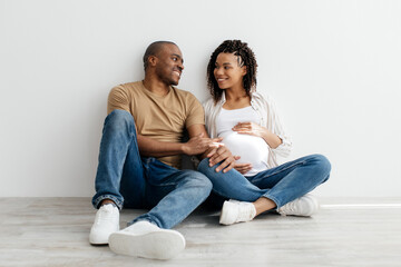 Portrait of romantic young black couple waiting for baby sitting on floor and looking at each other, loving african american pregnant woman and her husband ready to be parents, copy space