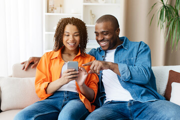 Beautiful Black Couple Using Mobile Phone And Embracing Together At Home, Happy African American Spouses Relaxing On Couch With Modern Smartphone, Browsing New App Or Shopping Online, Copy Space