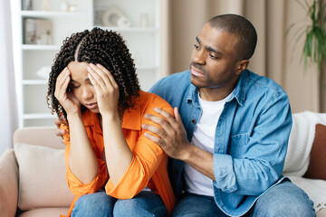 Compassionate black man comforting his upset wife at home, loving husband providing support and expressing empathy while sitting together on couch in living room interior, closeup shot