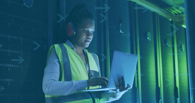 Mid-adult woman technician wearing vest examining racks in server room aisle, holding laptop