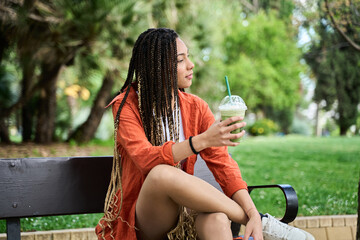 Young latina woman with braids enjoying a refreshing drink in the park