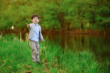 curious boy by serene lake holding a blade of grass, exploring lush nature in springtime. green background and tranquil water create peaceful outdoor scene capturing childhood wonder