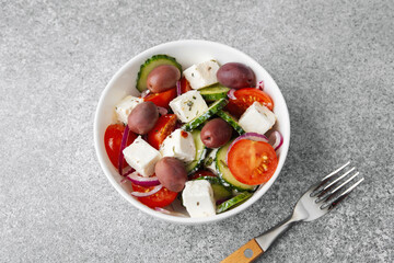 Greek salad with feta cheese olives cucumbers tomatoes onions in white bowl with fork on light grey background