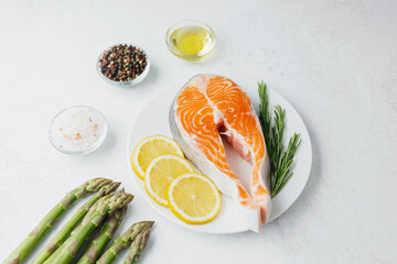 Raw salmon steak with lemon slices, rosemary, asparagus and spices on white plate over light background