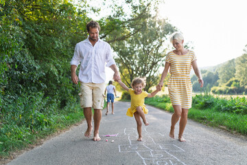Parents helping litthe girl with hopscotch during warm summer day.