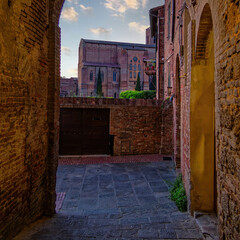 The Basilica Cateriniana di San Domenico in Siena, Tuscany, Italy, emerges beautifully through an old arched alley, framed by historic stone walls and bathed in soft natural light.