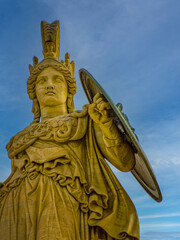 Marble statue of Athena, the ancient Greek goddess of wisdom and knowledge, with a helmet and a shield. The craftsmanship highlights carvings and drapery, against a backdrop of a sky with clouds.