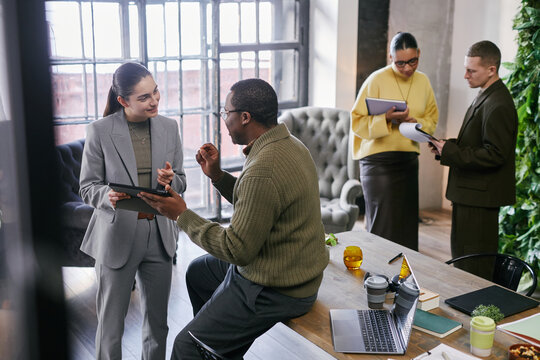 Multiethnic group of young adult and middle aged men and women discussing business strategy in modern office, using digital tablets and laptops, collaborating on project ideas