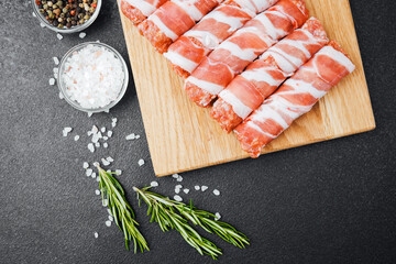 Raw meat rolls wrapped in bacon on wooden board with spices