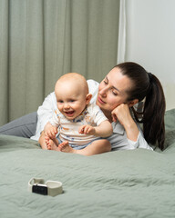 Smiling mother relaxes on bed while her curious one year old son crawls toward wristwatch. Candid...