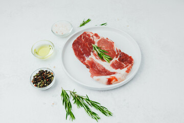 Uncooked pork steak on white plate with rosemary and pepper