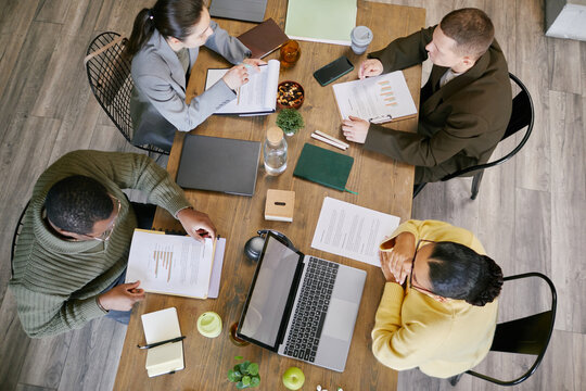 High angle shot of diverse group of young adult and middle aged men and women collaborating around table, reviewing business documents, using laptop and digital tablet, discussing financial charts
