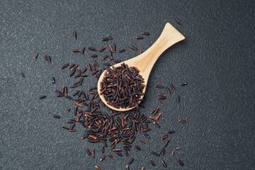 Wooden spoon filled with black rice and spilled grains on dark background