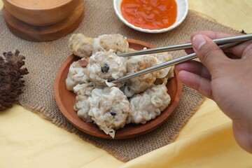 A hand holding chopsticks to pick up dimsum dumplings from a plate, with a side of dipping sauce...