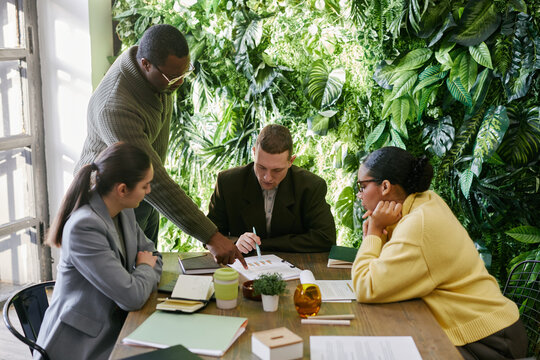 Middle aged Black man guiding multiethnic team pointing at documents while analyzing collaboratively business reports at desk in modern office with plant wall