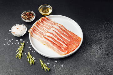 Raw bacon slices on white plate with pepper, salt and rosemary
