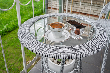 A cup of coffee, a smartphone on a glass table on a balcony.	
