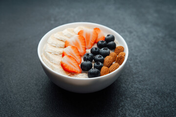 Oatmeal porridge with banana strawberry blueberry and almond toppings on dark background