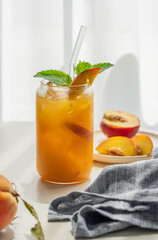 Peach drink or tea with ice, mint in a glass on a white table near window with fresh fruits