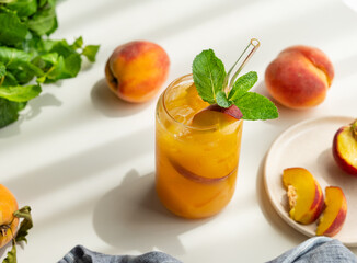 Peach drink or tea with ice, mint in a glass on a white table with fresh fruits and sunlight.