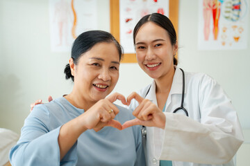 Asian woman Doctor and Elderly Woman Smiling Together Making heart shape with their hands in Clinic,symbolizing care,trust,strong doctor-patient relationships in compassionate healthcare environment.
