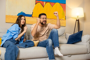 Excited couple sitting on modern sofa watching show or sports game on TV