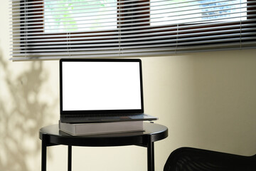Laptop with blank screen on top of books on a black round table near window blinds