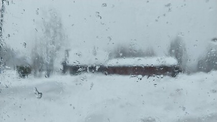 Blurred view of a snow-covered landscape through a frosty window, featuring a distant red building amidst heavy snowfall.