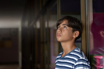 A contemplative teenage boy in a striped blue shirt looks upward indoors, standing near a glass wall with soft natural light. Mood of hope, thought, detention, lonely, sad, broken family and solitude.