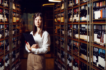 Professional young female sommelier standing in wine cellar between wine shelves with various wines, horizontal indoor portrait.