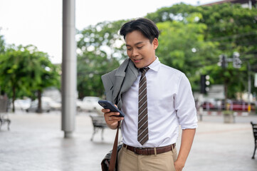 Smiling male office worker holding his phone with suit jacket draped on shoulder while standing in...