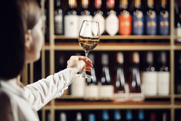 Vintner woman tasting white wine from a glass in cellar. Close up shot of female sommelier hand...