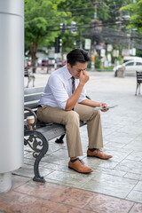 Male office worker massaging or rubbing his eyes while holding tablet sitting on steel bench in park