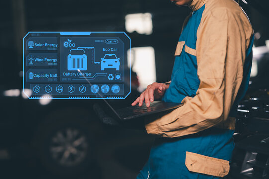 Technician in work overalls uses a laptop to monitor eco car battery charging via solar and wind energy on a futuristic digital interface.