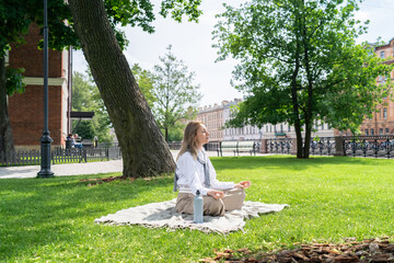 Calm tranquil woman in yoga lotus position meditating on grass in summer park alone, inner peace. Female resting in nature, recovering from daily stress, surrounded by greenery, mental wellbeing