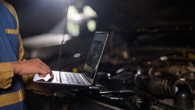 A technician uses a laptop to diagnose or repair a car engine in a dimly lit workshop.