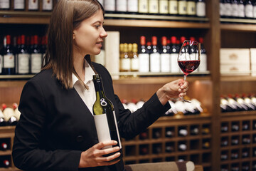Skilled sommelier tasting wine into glass standing in cellar over plenty bottle shelf background.