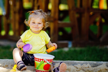 Cute toddler girl playing in sand on outdoor playground. Beautiful baby having fun on sunny warm summer sunny day. Happy healthy child with sand toys and in colorful fashion clothes.