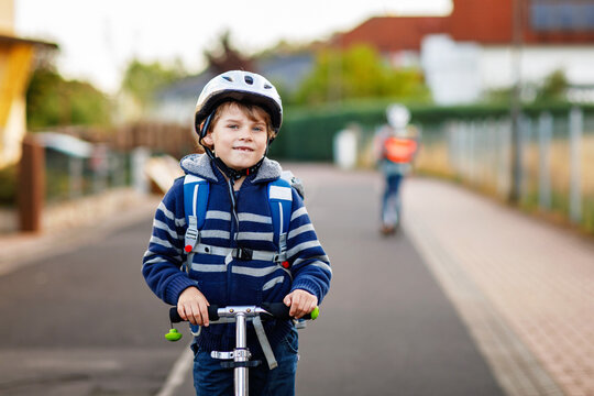 Active school kid boy in safety helmet riding with his scooter in the city with backpack on sunny day. Happy child in colorful clothes biking on way to school. - Powered by Adobe