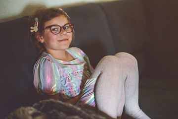 Little school girl with eyeglasses watching TV at home. Happy child enjoying a cozy moment indoors. Real life, real people, everyday childhood scene in a warm family environment.