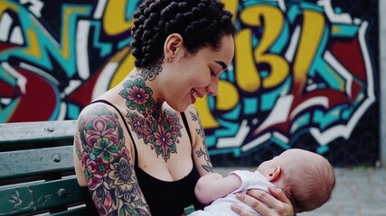 Tattooed young mother breastfeeding baby on bench near colorful graffiti wall