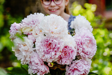 Close-up of little school girl holding a large bunch of blooming pink peonies in a garden. Happy child enjoying a spring or summer day outdoors. Real life, real people, natural childhood moment