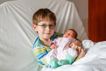 Happy boy with glasses holding his sleeping newborn sister in hospital bed. Real life, real people moment of sibling love, bonding, and family joy after birth.
