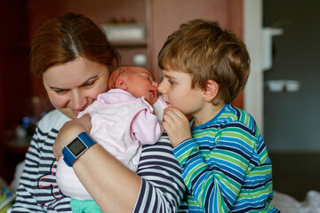 Happy little boy kissing his sleeping newborn sister in hospital bed. Mother holding baby girl on arm. Real life, real people moment of sibling love, bonding, and family tenderness