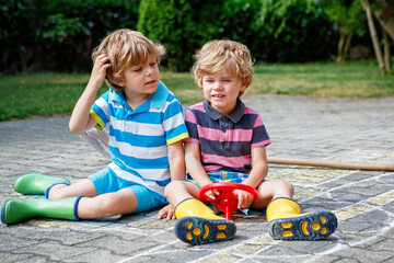 Two happy little boys sitting on pavement with colorful chalk drawing of a car. Siblings playing outdoors on sunny summer day. Kids wearing rubber boots, holding toy steering wheel.